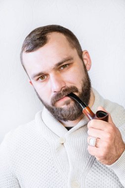 Hafif hırkalı, sakallı ve bıyıklı gülümseyen bir beyefendi pipo tutuyor ve sigara içiyor, şık, modaya uygun bir adam.