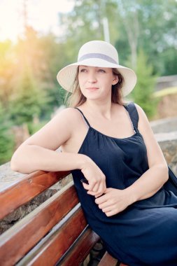 young woman on a bench in a hat looks into the distance at sunset