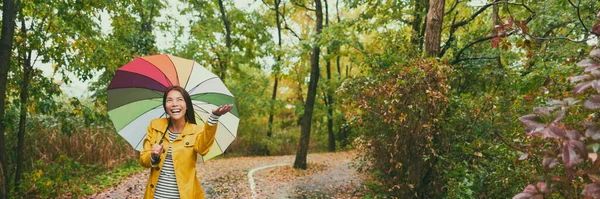 Mujer de otoño caminando bajo la lluvia bajo el paraguas. Chica asiática feliz disfrutando del bosque del parque al aire libre mirando hacia arriba alegre en el día lluvioso otoño vistiendo impermeable amarillo afuera en la naturaleza. Multiracial chica asiática —  Fotos de Stock