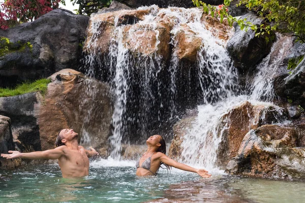 Pareja de naturaleza relajándose bajo cascada con los brazos abiertos en libertad. Personas disfrutando del agua que cae en la piscina natural en el fondo de la naturaleza tropical. Bienestar, salud y spa — Foto de Stock