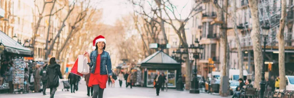 Weihnachtseinkäuferin, die mit Einkaufstaschen Geschenke kauft, trägt Weihnachtsmütze draußen auf der Straße. Transparent der sozialen Distanzierung des Konsumkonsums — Stockfoto