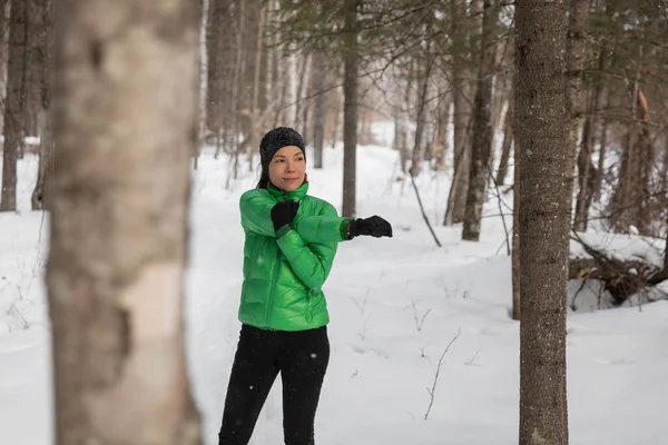 冬のフィットネス選手の女性は、屋外でスノートレイルを走る前に腕を伸ばして温暖化します。寒い天候の手袋、ヘッドバンド、ジャケットを身に着けているアジアのランナー — ストック写真