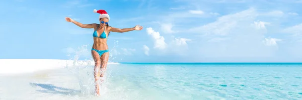 Christmas beach holiday travel banner panorama background for Christmas vacation fun. Bikini woman running carefree splashing water enjoying swim caribbean travel getaway with santa hat. — Stock Photo, Image