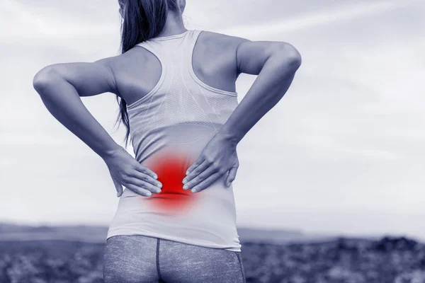 Травма спины в спорте. Девушка-спортсмен держит нижнюю часть спины от боли, массируя мышцы, касающиеся круга красной зоны, чтобы показать пораженный участок с синим фильтром медицинского цвета. — стоковое фото