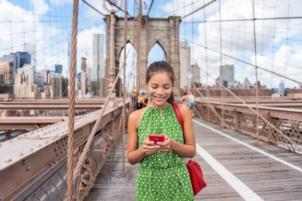 NYC resor telefon SMS flicka håller smartphone på Brooklyn bron i New York, Manhattan USA. Asiatisk kvinna telefon läsa eller använda sociala medier på sommaren. — Stockfoto