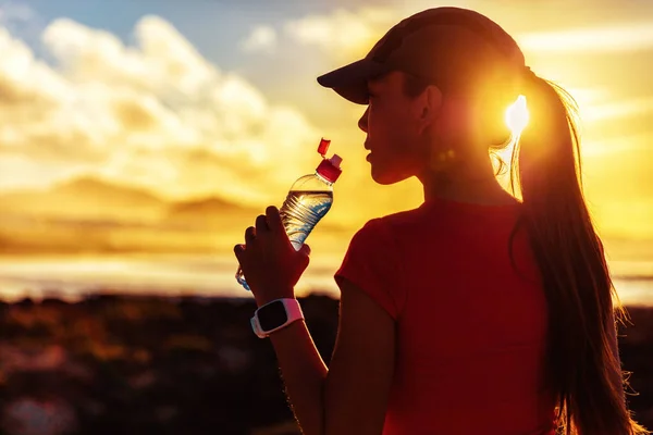 Mujer fitness beber agua de la botella de deportes en el entrenamiento de la tarde después de correr entrenamiento correr al aire libre al atardecer. Chica con gorra de correr silueta contra el sol bengala —  Fotos de Stock