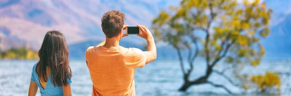 Travel lifestyle people tourists taking picture with phone on New Zealand Wanaka tree background Vacation couple banner panorama. — Stock Photo, Image