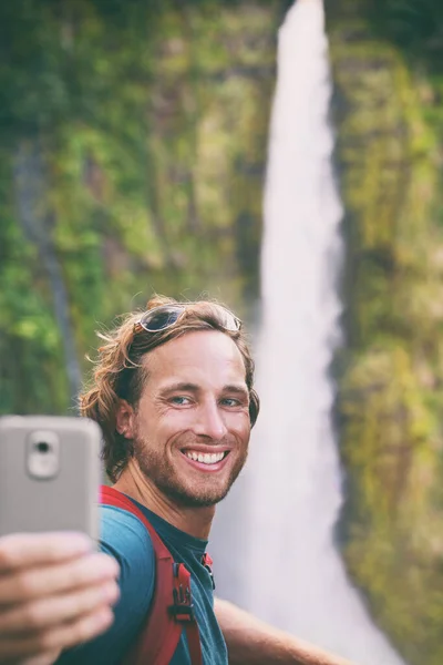 Selfie seyahat turisti Hawaii şelalesi manzarasında gezerken teknolojik akıllı telefon uygulaması ile fotoğraf çekiyor. Gülümseyen Kafkasyalı genç adam yaz tatili macerasında elinde telefonla gezintiye çıkıyor.. — Stok fotoğraf