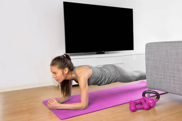 लिविंग रूम फिटनेस कसरत घर पर कोर व्यायाम करने के लिए प्लैंक व्यायाम करने वाली लड़की। युवा एशियाई महिला जिम जाने के बिना एक स्वस्थ जीवन शैली के हिस्से के रूप में टीवी के सामने मांसपेशियों को प्रशिक्षण दे रही है . — स्टॉक फ़ोटो, इमेज