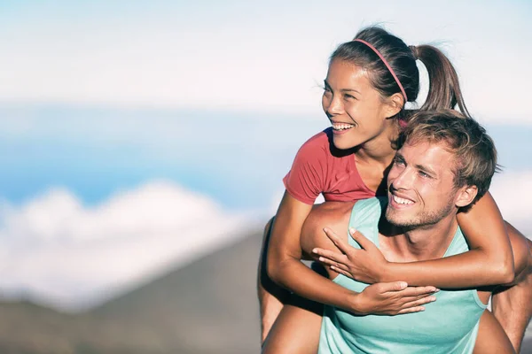 Casal feliz sorrindo em diversão viagem aventura, relacionamento interracial mulher asiática e homem caucasiano. Menina piggybacking abraçando namorado. Fitness pessoas apaixonadas juntas ao ar livre olhando para a natureza. — Fotografia de Stock