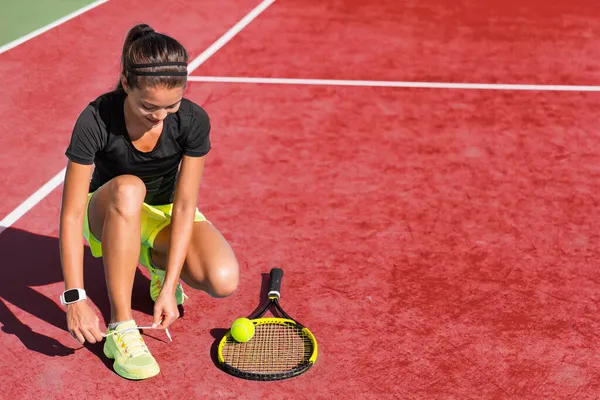 Sport fitness woman getting ready to play tennis on red clay court tying up shoe laces. Exercise summer training fit girl motivation. Racket and tennis ball on background.