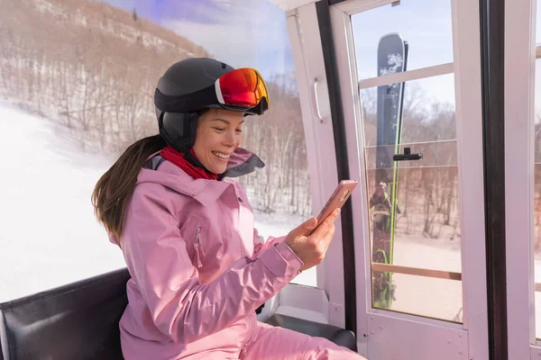 滑雪度假-女滑雪者使用电话应用程序在贡多拉滑雪电梯。女孩笑着看着手机，穿着滑雪服，头戴头盔和护目镜。滑雪冬季活动度假概念 — 图库照片
