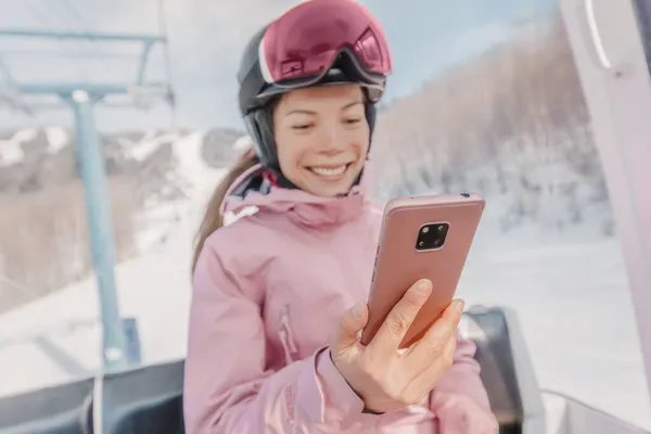 Telefon na nartach - kobieta narciarz za pomocą aplikacji telefonicznej w gondoli wyciąg narciarski. Dziewczyna uśmiechnięta patrząc na mobilny smartfon w ubraniu narciarskim, kasku i goglach. Koncepcja wakacyjnej aktywności narciarskiej — Zdjęcie stockowe
