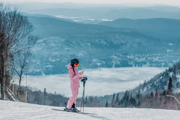 Telefone em férias de esqui - Mulher esquiador usando aplicativo de telefone na pista de esqui inclinação na paisagem incrível natureza inverno. Menina olhando para smartphone móvel vestindo roupas de esqui incrível, capacete e óculos — Fotografia de Stock