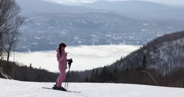 스키 여행에 관한 전화 - 스키 트레일 경사면에서 핸드폰 앱을 사용하는 여성 스키어 놀라운 겨울 풍경. 스키멋진 옷을 입고 헬멧을 쓰고 고글을 쓰고 휴대폰을 보고 있는 소녀 — 비디오