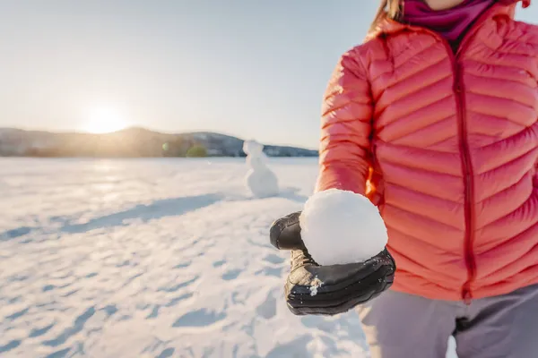 Invierno al aire libre concepto de estilo de vida activo. Niña sosteniendo bola de nieve en el día nevado con muñeco de nieve en el fondo en el lago congelado en el hermoso atardecer de invierno —  Fotos de Stock