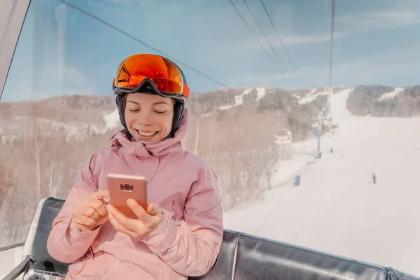 Wakacje narciarskie - kobieta narciarz za pomocą aplikacji telefonicznej w gondoli wyciąg narciarski. Dziewczyna uśmiechnięta patrząc na mobilny smartfon w ubraniu narciarskim, kasku i goglach. Aktywność narciarska zima koncepcji wakacje — Zdjęcie stockowe