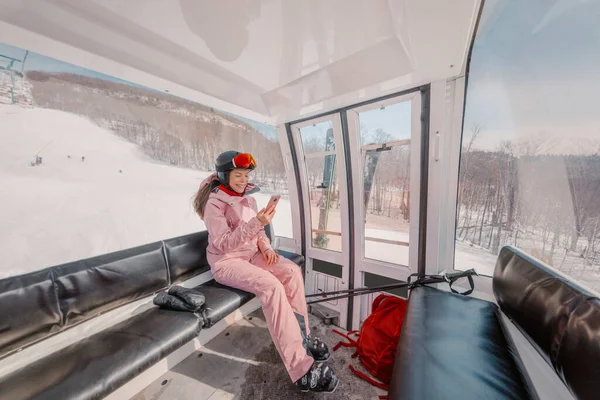 Kayak tatillerinde teleferikle teleferik kullanan kadın kayakçı. Havalı kayak kıyafetleri, miğfer ve gözlüklerle cep telefonu kullanarak gülümseyen bir kız. Kış tatili etkinliği kavramı — Stok fotoğraf