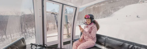 Ski Winter Holidays - skier using phone app in gondola ski lift. Woman smiling looking at mobile smartphone wearing ski clothing, helmet and goggles. Ski winter activity concept panoramic banner — Foto Stock