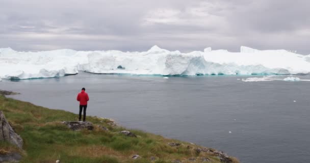 Travel adventure in Arctic landscape nature with icebergs - tourist person looking at view of Greenland icefjord - aerial video. Man by ice and iceberg, Ilulissat Icefjord — Vídeo de Stock