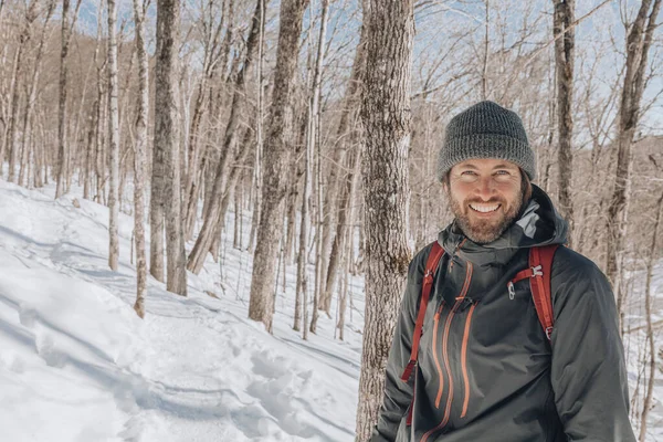 Winterwandelportret van een vrolijke man glimlachend kijkend naar de camera in het sneeuwwoud berglandschap. Mannelijke wandelaar wandelen met rugzak in koud weer natuur landschap — Stockfoto