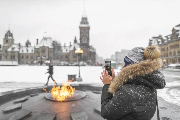 Ottawa travel tourist woman taking photo with phone of Canadian Parliament in Ontario, Canada. Snowing landscape and centennial flame that do not freeze in winter — Stockfoto