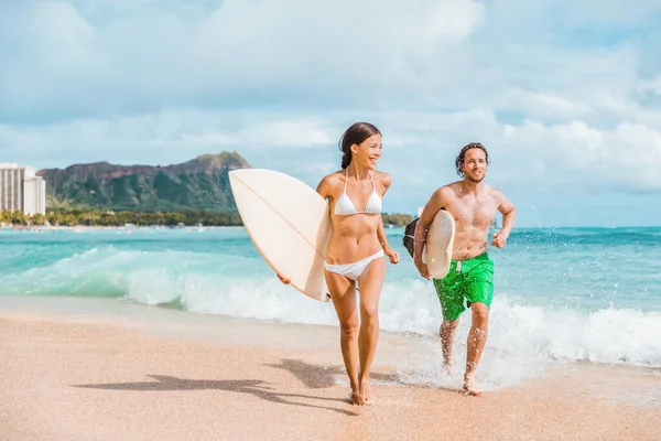 Hawaii surfing couple surfers in Waikiki beach, Honolulu Asian girl and Caucasian guy surfer carrying surfboards running out of water. Surf lifestyle Oahu island. USA travel. Fun vacation activity — Fotografia de Stock