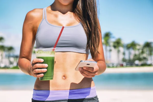 Healthy green smoothie juice drink online order on phone app fit athlete girl drinking detox cleanse morning breakfast on beach. Sport woman outside in summer sun with diet food — Stockfoto