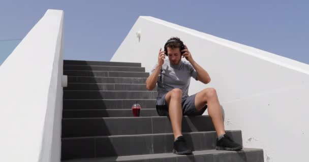 Hombre escuchando música, audiolibro o podcast aplicación de teléfono móvil con auriculares sentados en las escaleras. Persona de estilo de vida saludable de la ciudad utilizando el teléfono inteligente en el descanso con batido de jugo de remolacha roja de la mañana — Vídeos de Stock