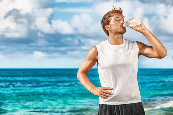 Sports drink fitness man drinking water botlle during outdoor exercise workout on beach. Dehydrated athlete runner after run sweating in summer heat exercising cardio — Foto de Stock