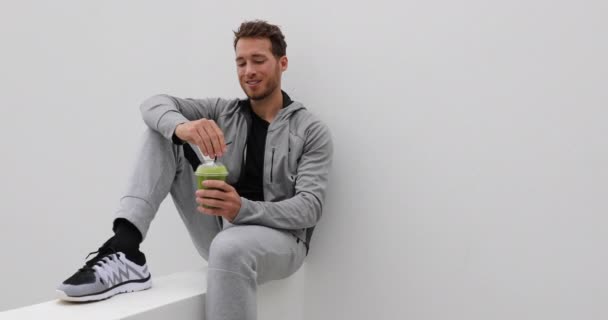 Hombre sano bebiendo batido verde en la terraza de casa. Deporte atleta con espinacas jugo prensado en frío copa de plástico bebida relajante al aire libre en pantalones de chándal y ropa deportiva casual — Vídeos de Stock