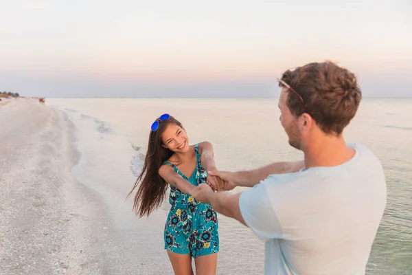 Happy Asian couple of lovers dancing in love on sunset beach. Smiling biracial woman holding hands of boyfriend during honeymoon travel vacation — Stock Photo, Image