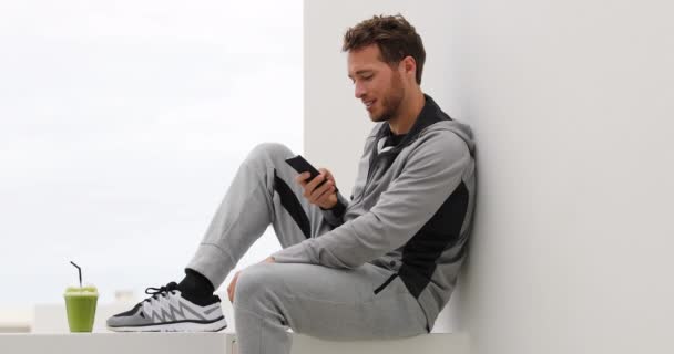 Teléfono. Joven hombre usando teléfono inteligente viendo vídeo en línea o mensajes de texto SMS mensajes. Hombre de fitness utilizando la aplicación del teléfono en el descanso corriendo relajarse al aire libre. Smoothie verde y pantalones de chándal estilo de vida activo saludable — Vídeos de Stock