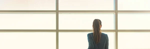 Ofis binasının penceresinden bakan iş kadını dalgın görünüyor. Arkadan iş adamı, sabah güneşine karşı siluet, kariyer hedefini ya da hayatı düşünüyor. Panorama Panoraması. — Stok fotoğraf
