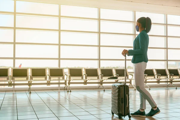 Coronavirus travel tourist wearing face mask in airport terminal watching windows with luggage. Unrecognizable business woman walking in lounge boarding gate at departure. Pandemic vacation lifestyle. — Stock Photo, Image