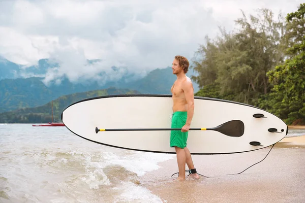 Paddleboard watersport fit man carrying stand up paddle board going exercising on ocean at Puu Poa beach, Hanalei Bay, Kauai, Hawaii, USA. Hawái viaje deporte atleta ejercicio — Foto de Stock