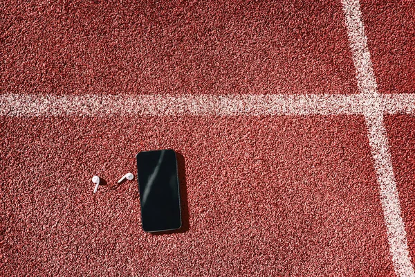 Fones de ouvido sem fio em faixas vermelhas do estádio de atletismo. Vista superior do fundo do telefone celular e fones de ouvido para o conceito de atleta fitness — Fotografia de Stock