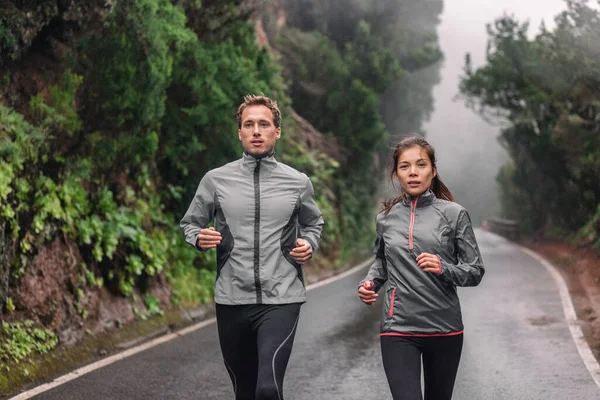 Correndo casal no parque molhado trilha correndo na chuva usando jaquetas sportswear em tempo frio. Mulher asiática, homem branco atletas formação cardio — Fotografia de Stock