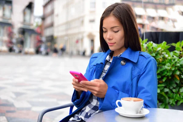 Café mujer mensajes de texto SMS en aplicación de teléfono beber café capuchino en terrasse calle al aire libre en la ciudad europea. Europa viaje estilo de vida. Mujer de negocios asiática en gabardina azul —  Fotos de Stock