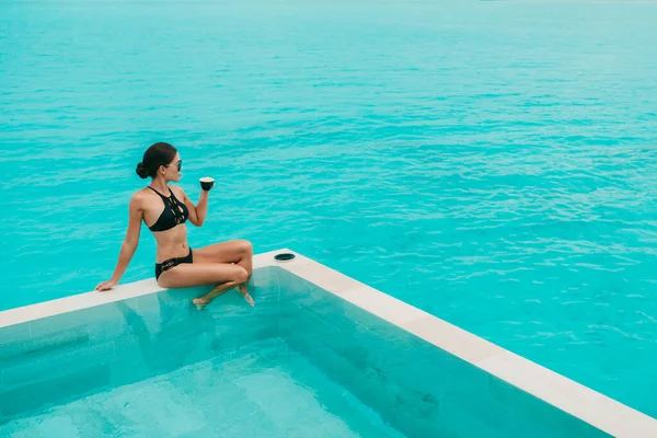 Mujer tomando café en la piscina del hotel de lujo con vista al océano turquesa y piscina infinita sobre el agua. Escapada al paraíso. Modelo de moda en bikini de lujo con estilo. — Foto de Stock