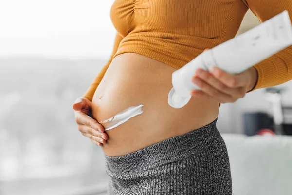 Zwangerschapsverzorging. Zwangere vrouw die crème op de maag aanbrengt voor striae. hydraterende buik close-up van handen en lotion fles thuis — Stockfoto