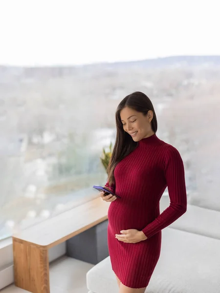 Asyalı hamile bir kadın cep telefonlu hamilelik uygulamasını doğum öncesi kursu için kullanıyor. Dikey olarak duran kırmızı elbiseli güzel bir model. — Stok fotoğraf