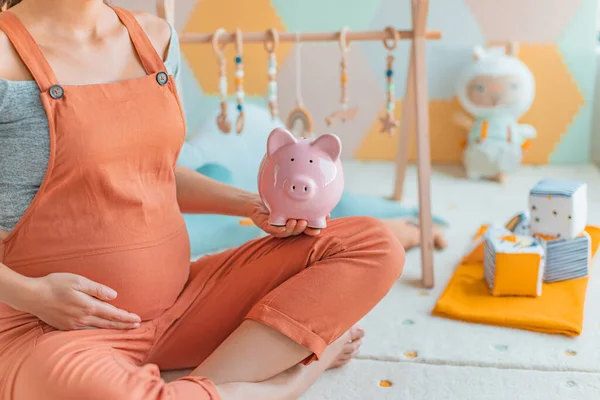 Pregnancy family planning budget. Cost of having a child. Pregnant woman holding piggy bank shopping newborn toys and nursery decor with savings. Maternity leave benefits