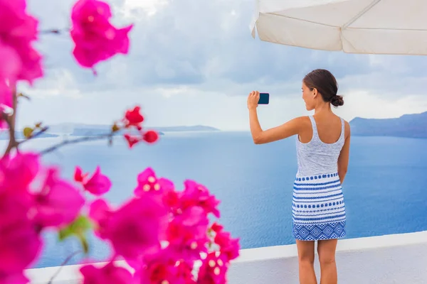 Reizen toeristische vrouw het nemen van foto 's met de telefoon van Santorini Middellandse Zee uitzicht vanaf de kust van grieks eiland. Europa Griekenland bestemming meisje sightseeing Oia in het voorjaar met roze bloemen voorgrond — Stockfoto