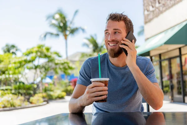 Man belt met behulp van de telefoon het maken van een gesprek drinken van koude koffie drinken zitten op cafe terras met smartphone praten op mobiele smartphone in de zomer. Knap jong casual man glimlachen gelukkig buiten Rechtenvrije Stockafbeeldingen