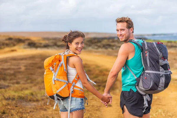 Šťastný mladý pár turistů v lásce drží za ruce chůze na trek túru s batohy. Turisté na turistické stezce přírody cíl cesty s úsměvem šťastný. Mezirasový vztah — Stock fotografie