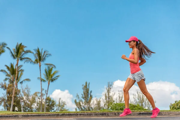 Felice corsa ragazza asiatica jogging nel parco il giorno d'estate indossando berretto rosa per la protezione solare. Corridore donna formazione cardio sul marciapiede. Profilo vista corpo intero dell'atleta — Foto Stock