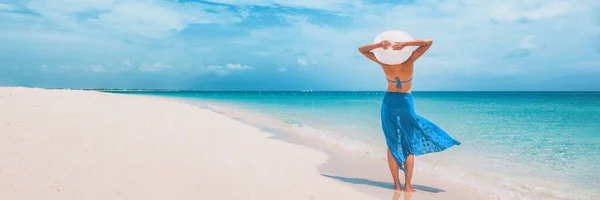 Vacaciones en la playa mujer feliz caminando en vacaciones de verano Vacaciones en el Caribe con los brazos detrás de la cabeza con sombrero de sol y falda sarong. Fondo de la bandera panorámica del océano. Elegante turista dama Fotos De Stock