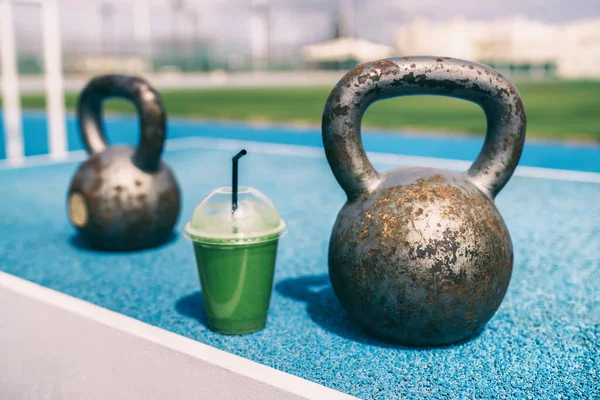 Zdrowie i fitness zielony smoothie detox drink na siłowni z obciążnikami kettlebells w centrum fitness na świeżym powietrzu treningu. Plastikowa filiżanka soku warzywnego śniadanie rano obok sprzętu wagi kettlebell — Zdjęcie stockowe