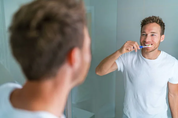 Brushing teeth man looking in mirror of home bathroom using toothbrush in morning routine for clean dental oral care — Stock Photo, Image